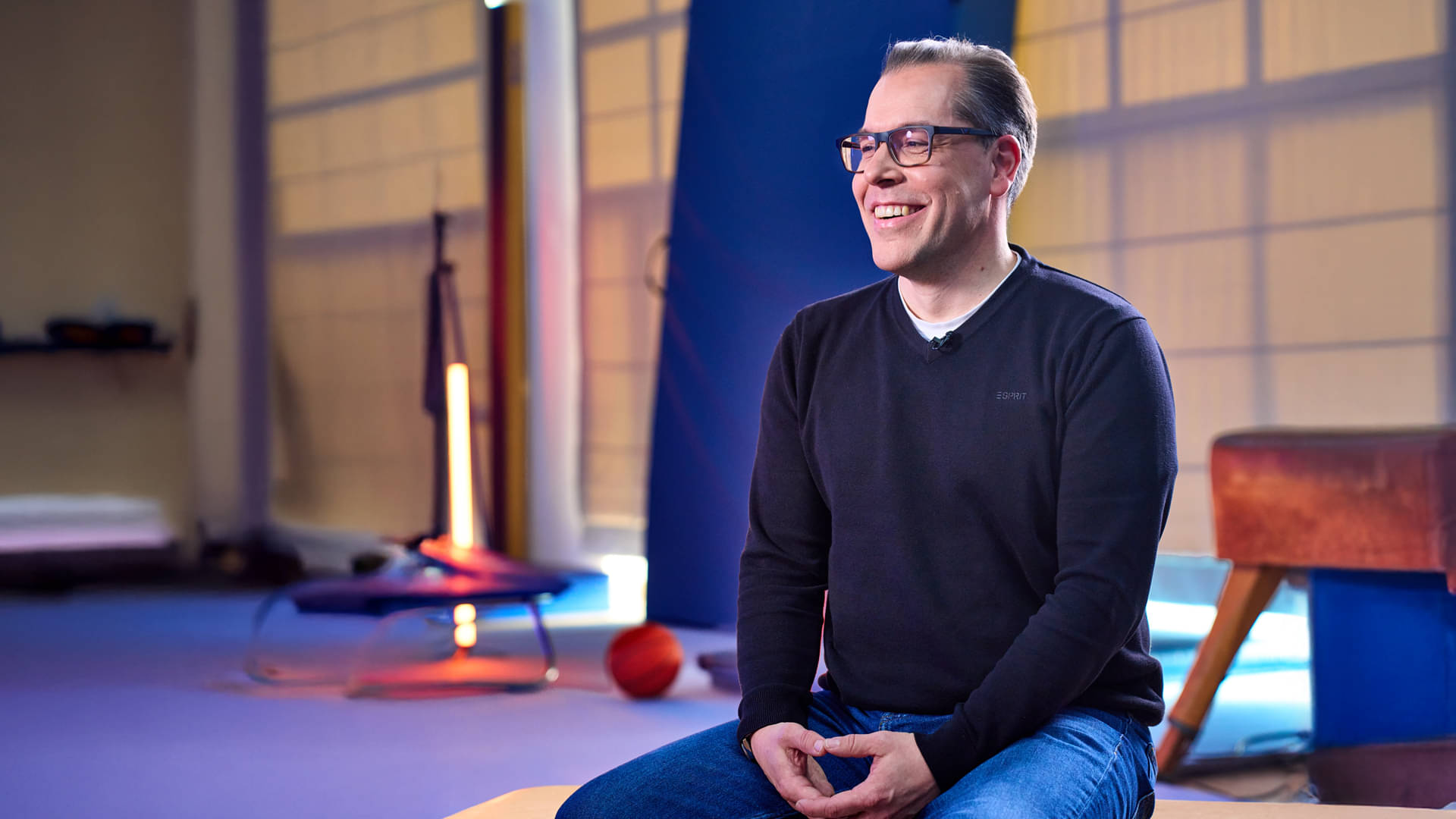 gefüllter_hocker, lockerer_sweater, brille, sitting_interview, studio_setting, Mann, Brille, Pullover, Jeans, Studio, 